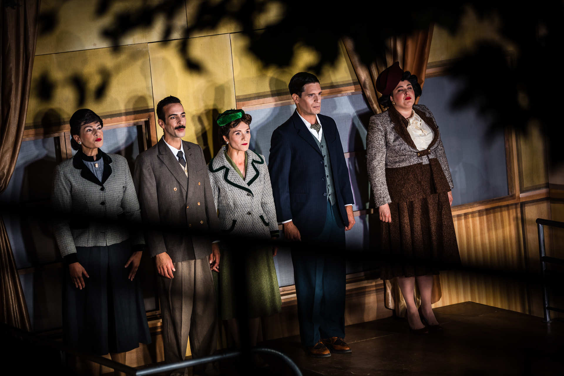 Cinco atores, três mulheres e dois homens, estão em palco. Conseguimos vê-los através de alguma vegetação, que aparece desfocada no campo da imagem. Estão vestido com roupa típica da década de 1950.
