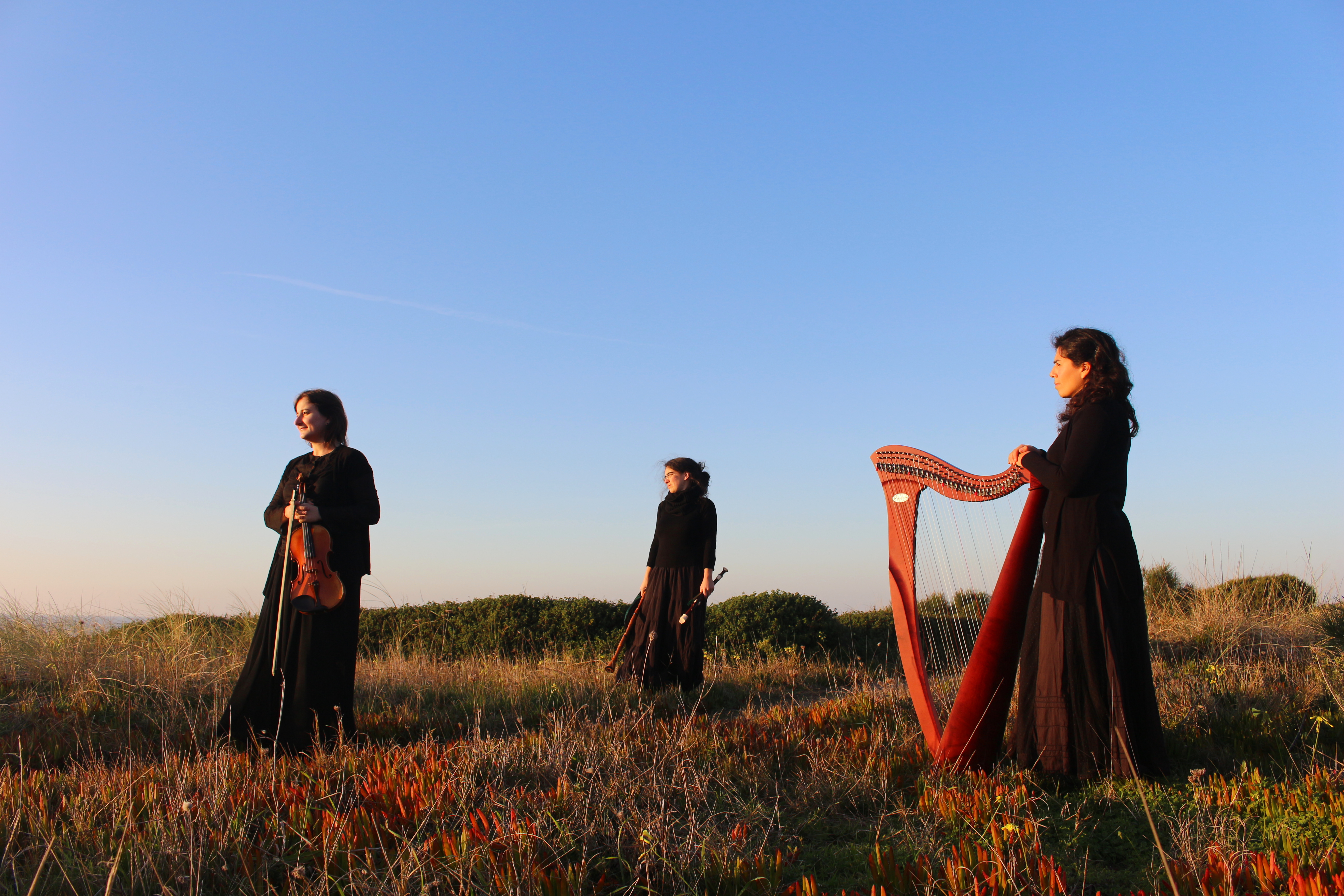 Três mulheres estão distribuídas ao longo de uma duna. As três estão vestidas de preto. A da esquerda tem um violino na mão, a do meio segura alguns instrumentos de sopro e a da direita tem uma harpa. A duna está coberta de vegetação, alguma verde, outra laranja e seca do sol. O céu está azul e límpido.