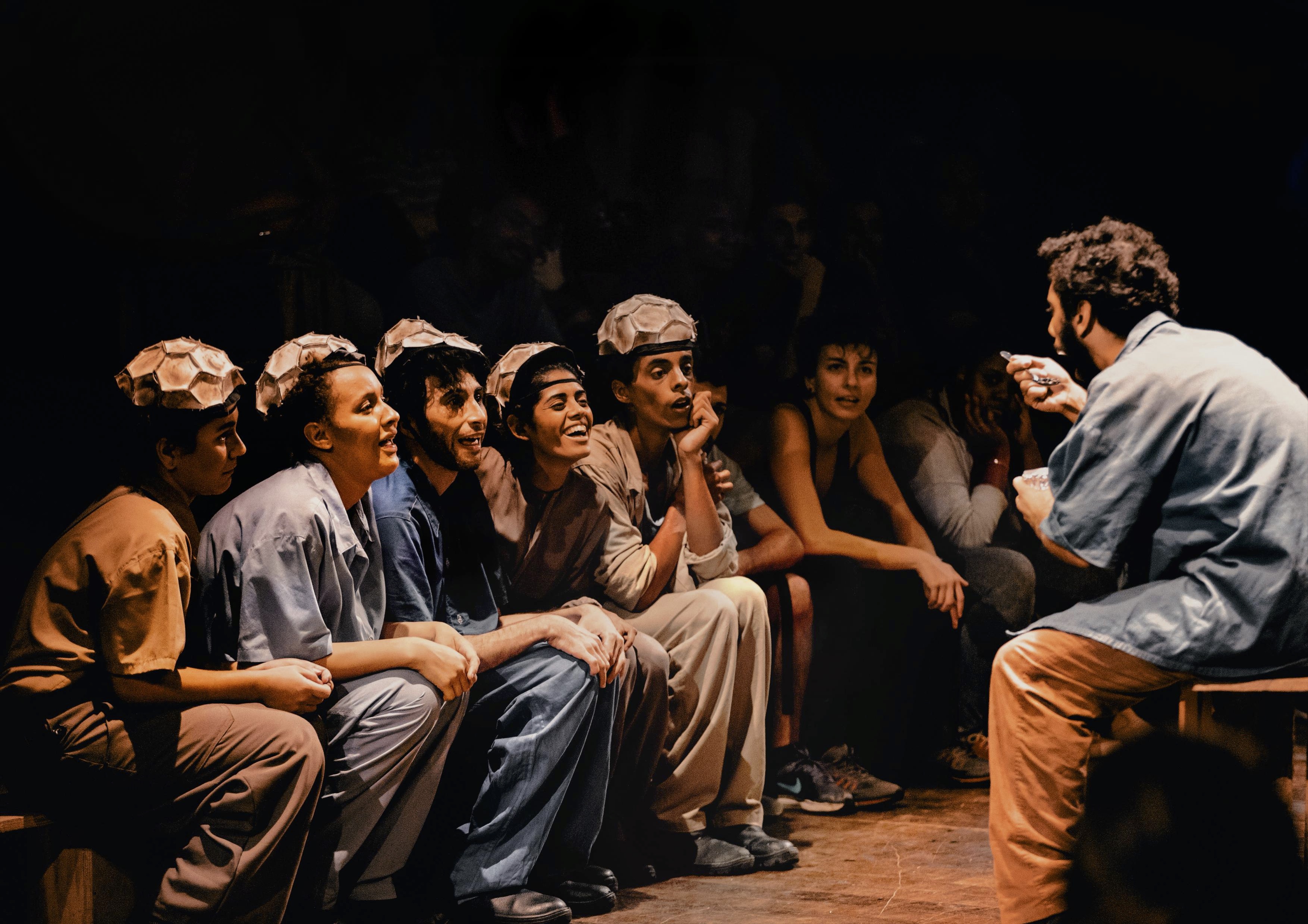 A imagem mostra uma cena de "Revolução na América do Sul" com um grupo de jovens atores e atrizes sentados em um banco, todos olhando atentamente para uma pessoa à direita que está a gesticular com as mãos. Os atores estão usando roupas simples e uniformes de cores neutras, como castanho e azul, e todos usam chapéus feitos de material que se assemelha a embalagens de caixas de ovos. A iluminação é quente e direcionada, destacando os rostos dos atores e suas expressões de concentração e curiosidade, enquanto o fundo está mais escuro, sugerindo um ambiente intimista. A cena transmite um momento de engajamento e emoção em uma performance teatral.