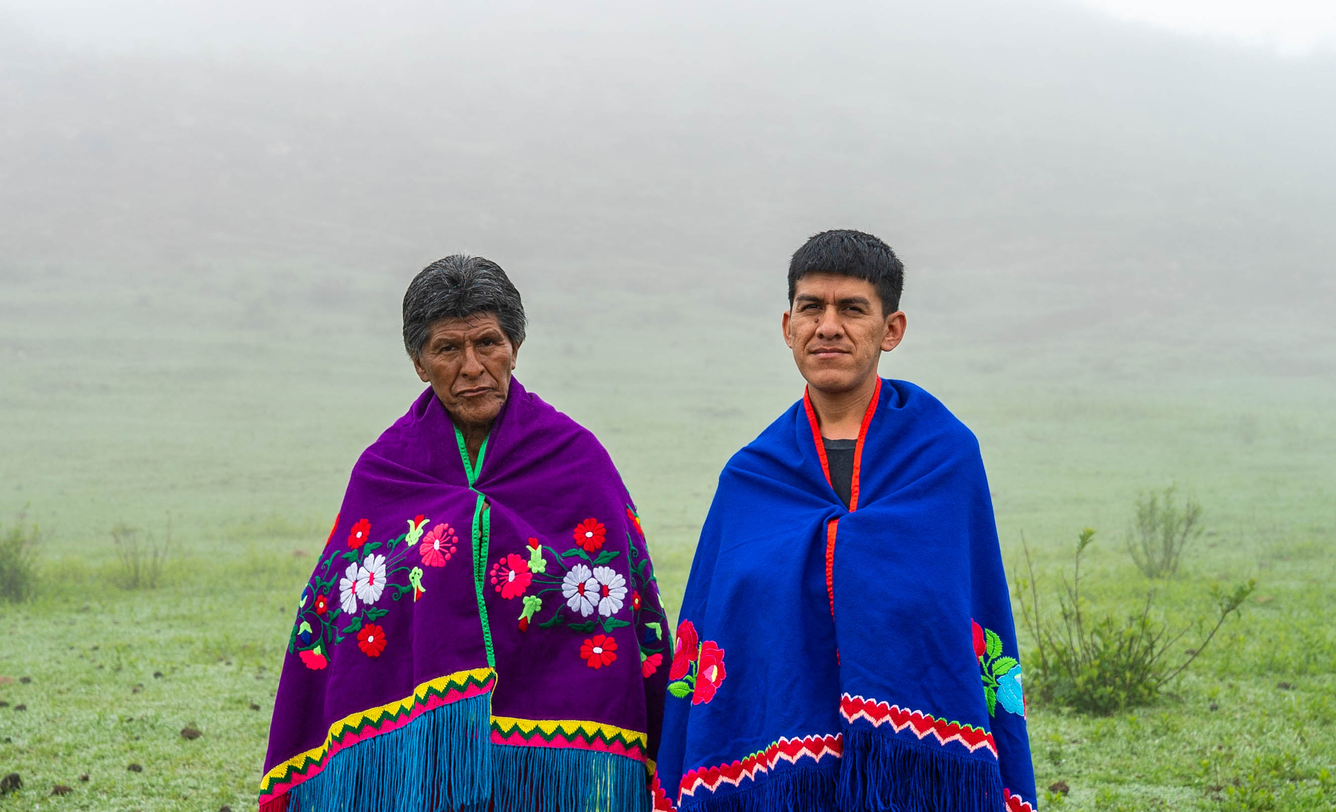 A imagem mostra dois homens indígenas em pé, lado a lado com um campo verde e com neblina ao fundo. Ambos estão usando cobertores coloridos e ornamentados, típicos de algumas culturas indígenas da América Latina. O homem à esquerda, mais velho, usa um cobertor roxo vibrante com flores bordadas em vermelho, rosa e branco, além de detalhes em verde. O homem à direita, mais jovem, veste um cobertor azul-escuro com bordados de flores em cores similares. A expressão deles é séria, e a paisagem ao fundo é enevoada, criando uma atmosfera serena e contemplativa.